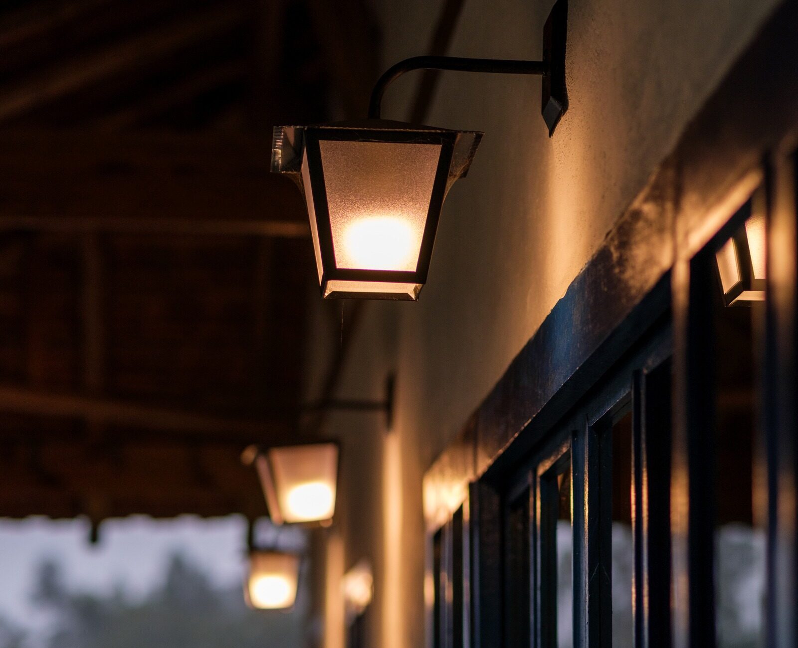 Warmly lit exterior lamps line a building's wooden overhang, casting soft light on a dark glass window with blurred greenery in the background.