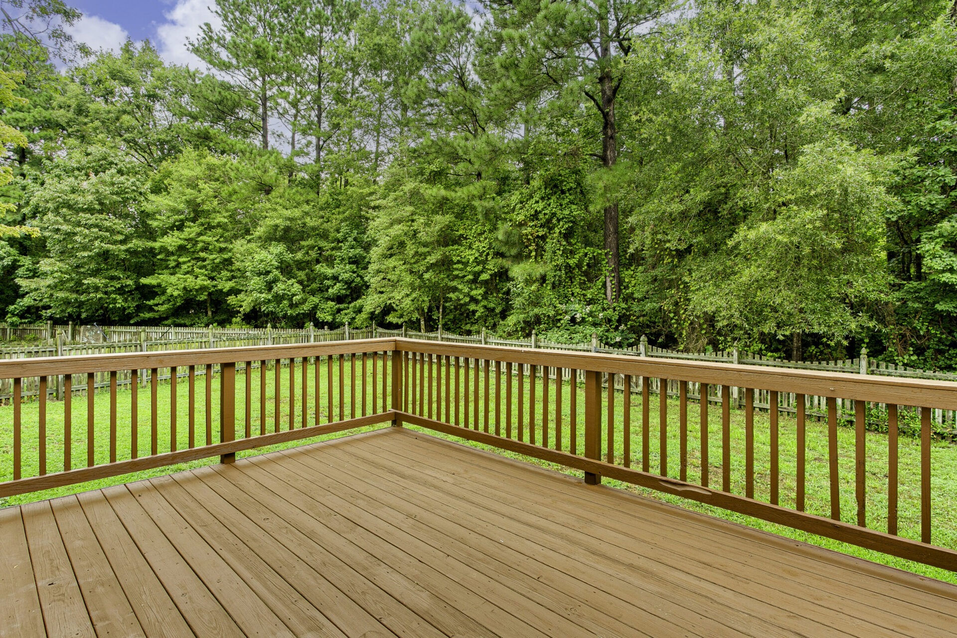 A wooden deck overlooks a lush, green backyard with a fence and dense tree line under a partly cloudy sky. Peaceful and inviting.