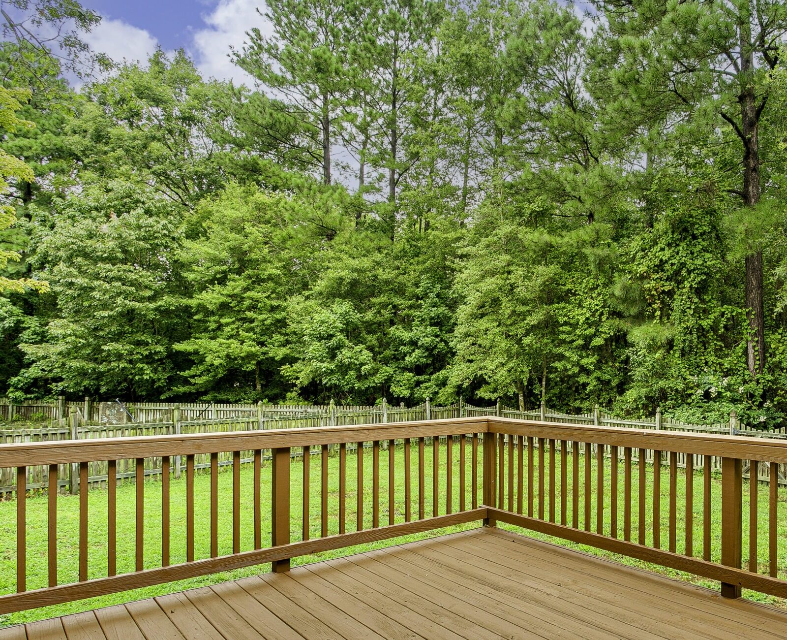 A wooden deck overlooks a lush, green backyard with a fence and dense tree line under a partly cloudy sky. Peaceful and inviting.