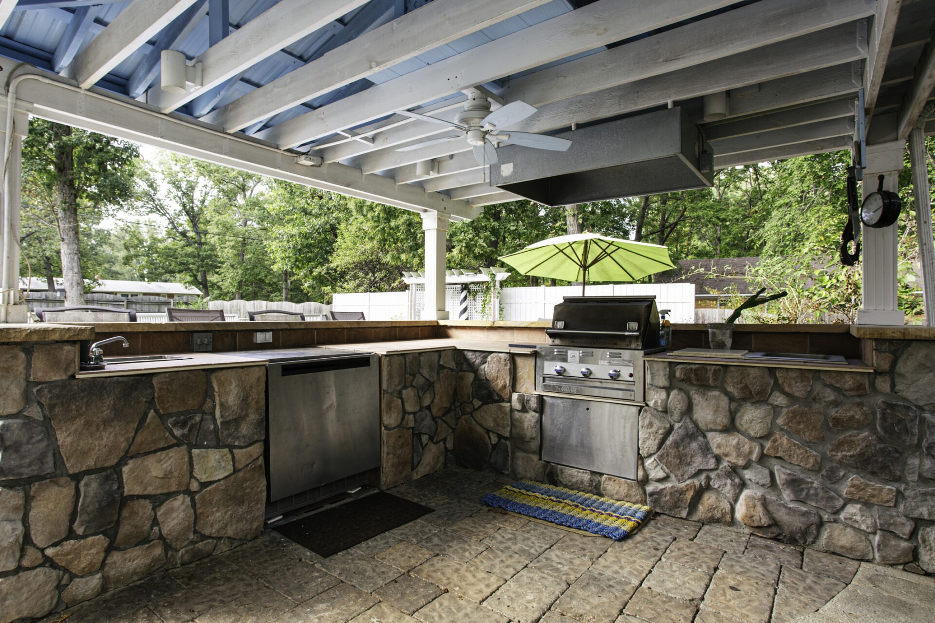 Outdoor kitchen with stone countertops, stainless steel grill, and ceiling fan. Surrounded by greenery and lounge chairs, under a wooden pergola.