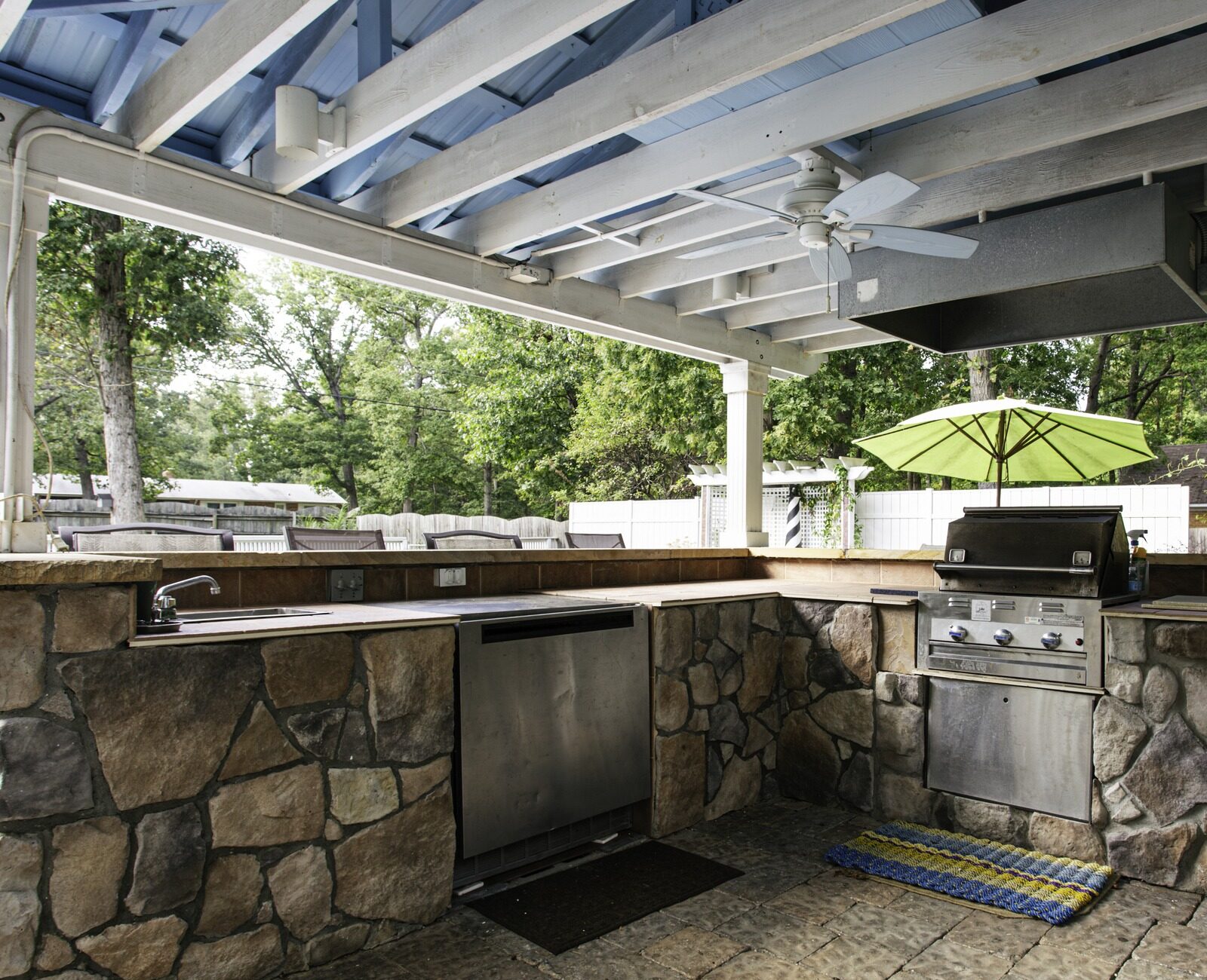 Outdoor kitchen with stone countertops, stainless steel grill, and ceiling fan. Surrounded by greenery and lounge chairs, under a wooden pergola.