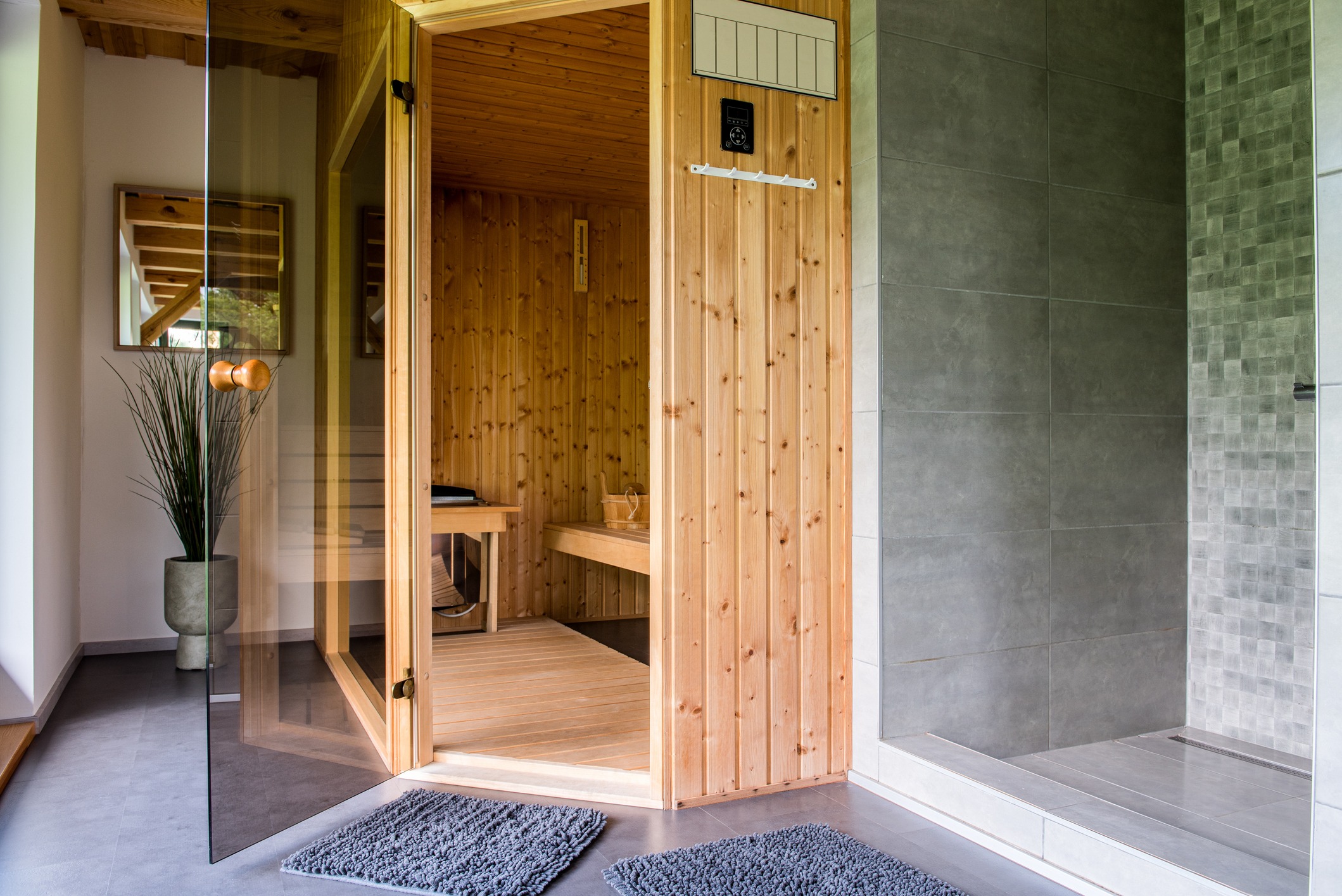 Cozy home sauna with wooden interior, open glass door, adjacent shower, modern gray tiles, and decorative plant in a stylish pot.