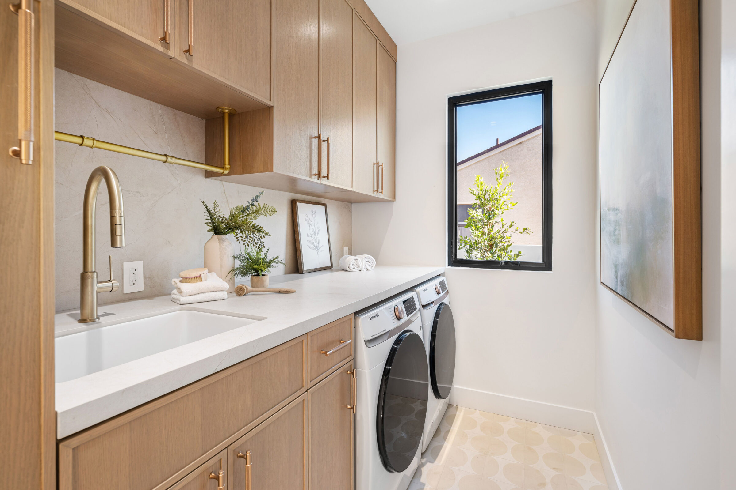 Modern laundry room with wooden cabinets, sink, and Samsung washer and dryer. Decor includes plants and framed picture near a bright window.