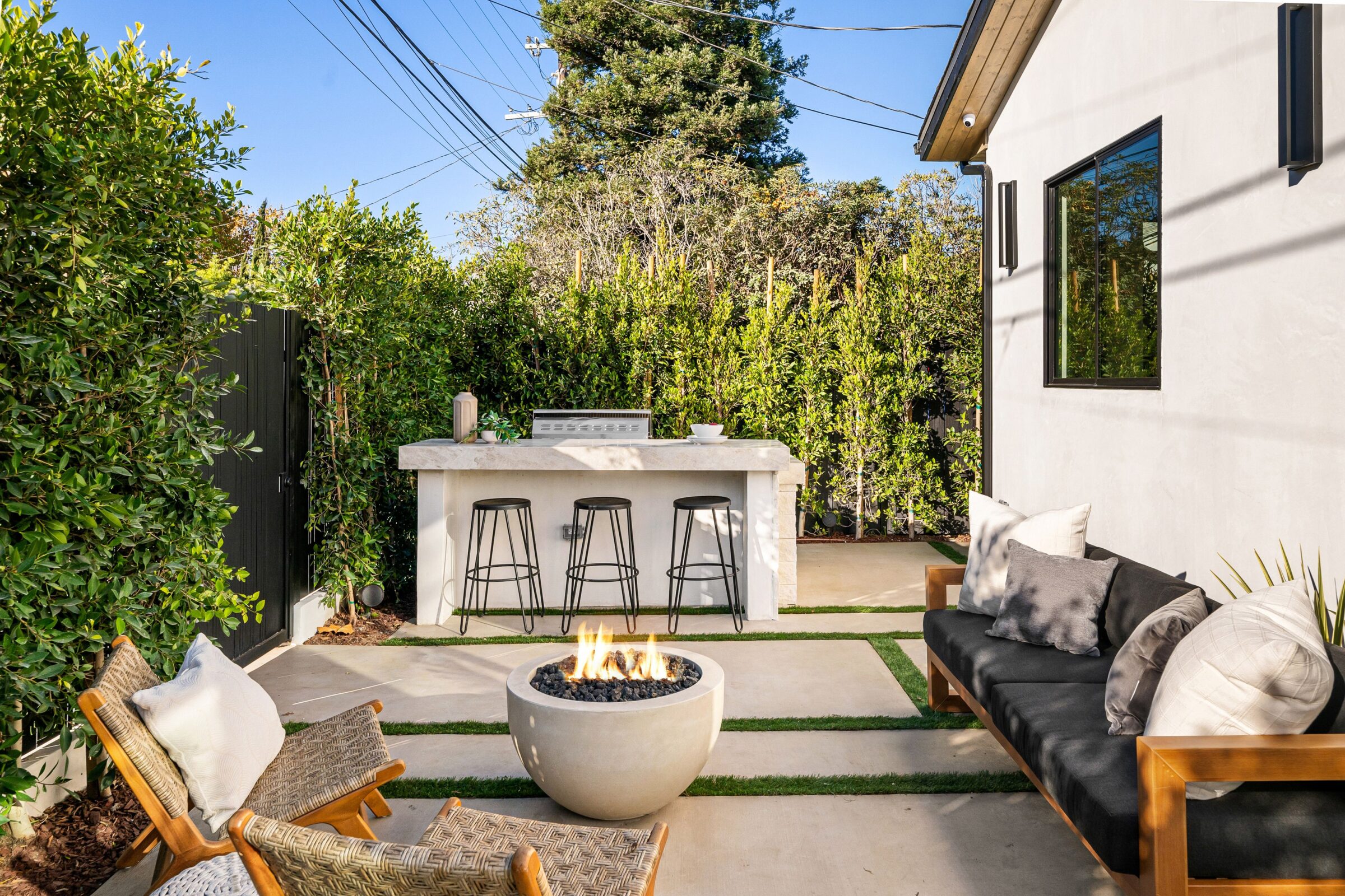 Modern outdoor patio with a fire pit, seating area, bar stools, lush greenery, and a sleek barbecue grill under clear blue sky.