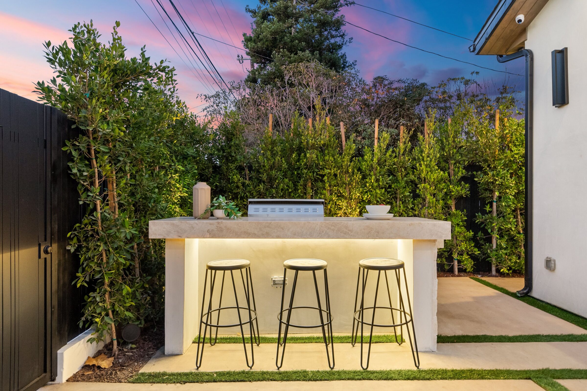 Outdoor bar setup with three stools, stone counter, plants behind, and evening sky in the background. Decorative item and bowl on counter.