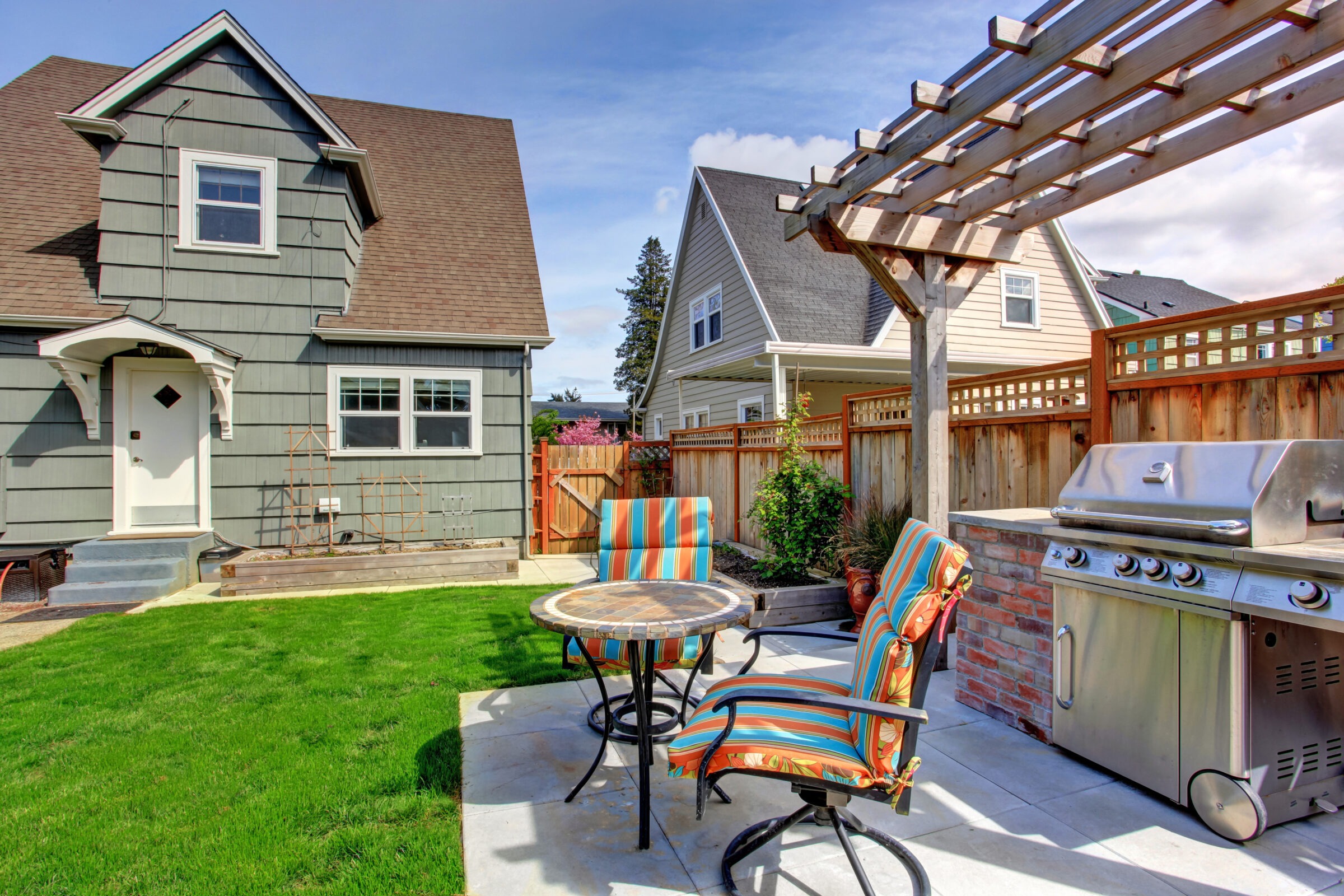 A suburban backyard features a gray house, patio set with striped chairs, pergola, grill, lush lawn, and wooden fence under a clear sky.
