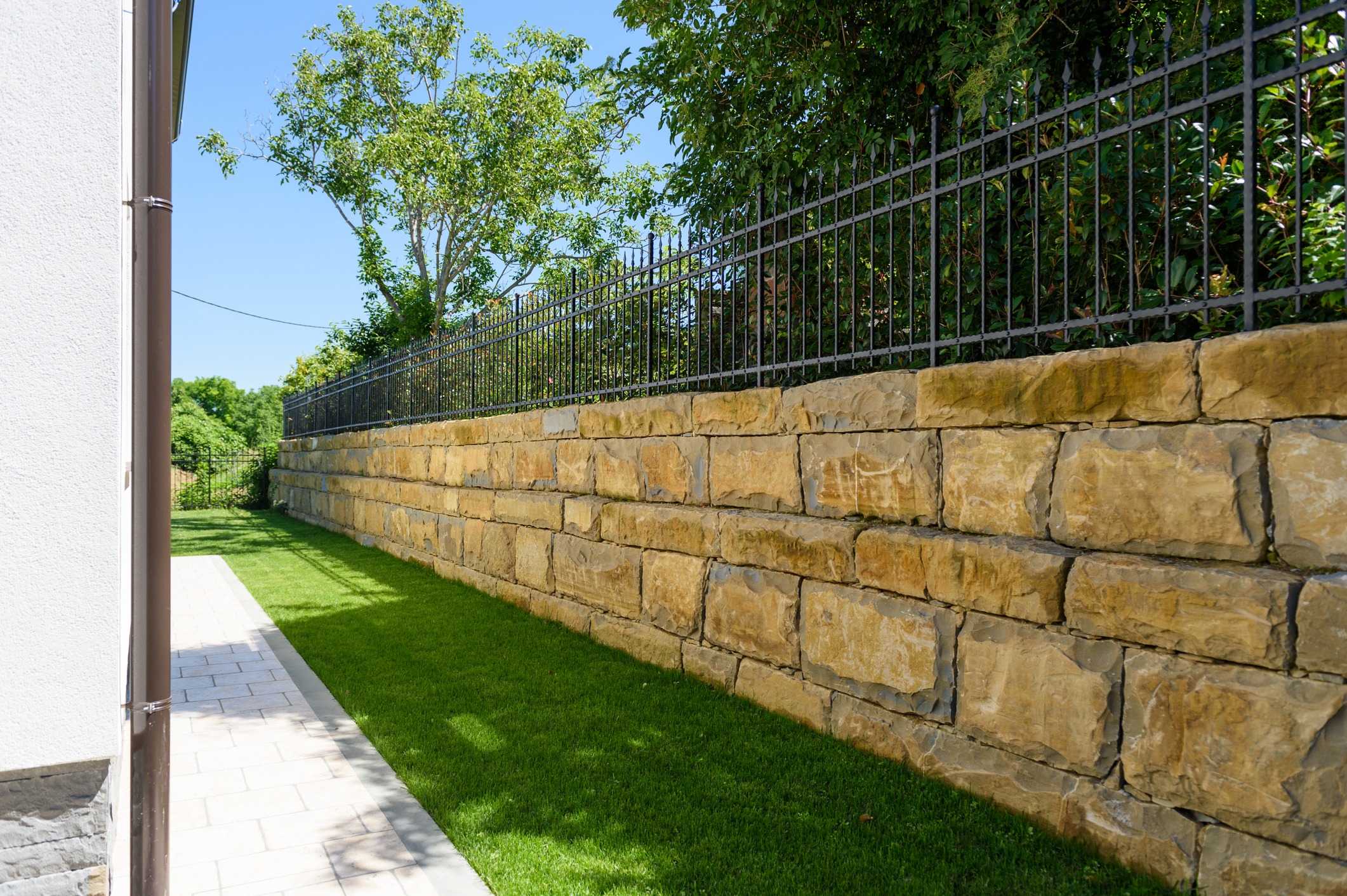 A stone wall with an iron fence borders a lush green lawn, adjacent to a white building, under clear blue sky.