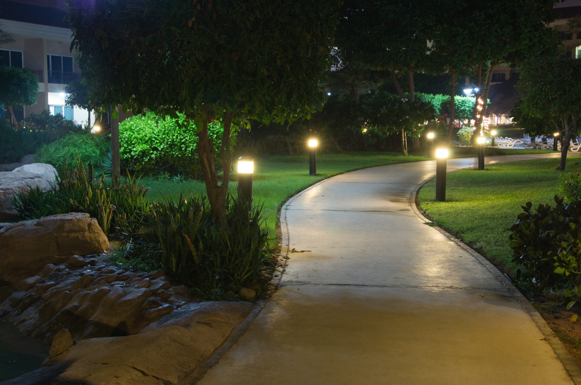 Illuminated garden path at night, surrounded by lush greenery and rocks, with buildings visible in the background, creating a serene atmosphere.