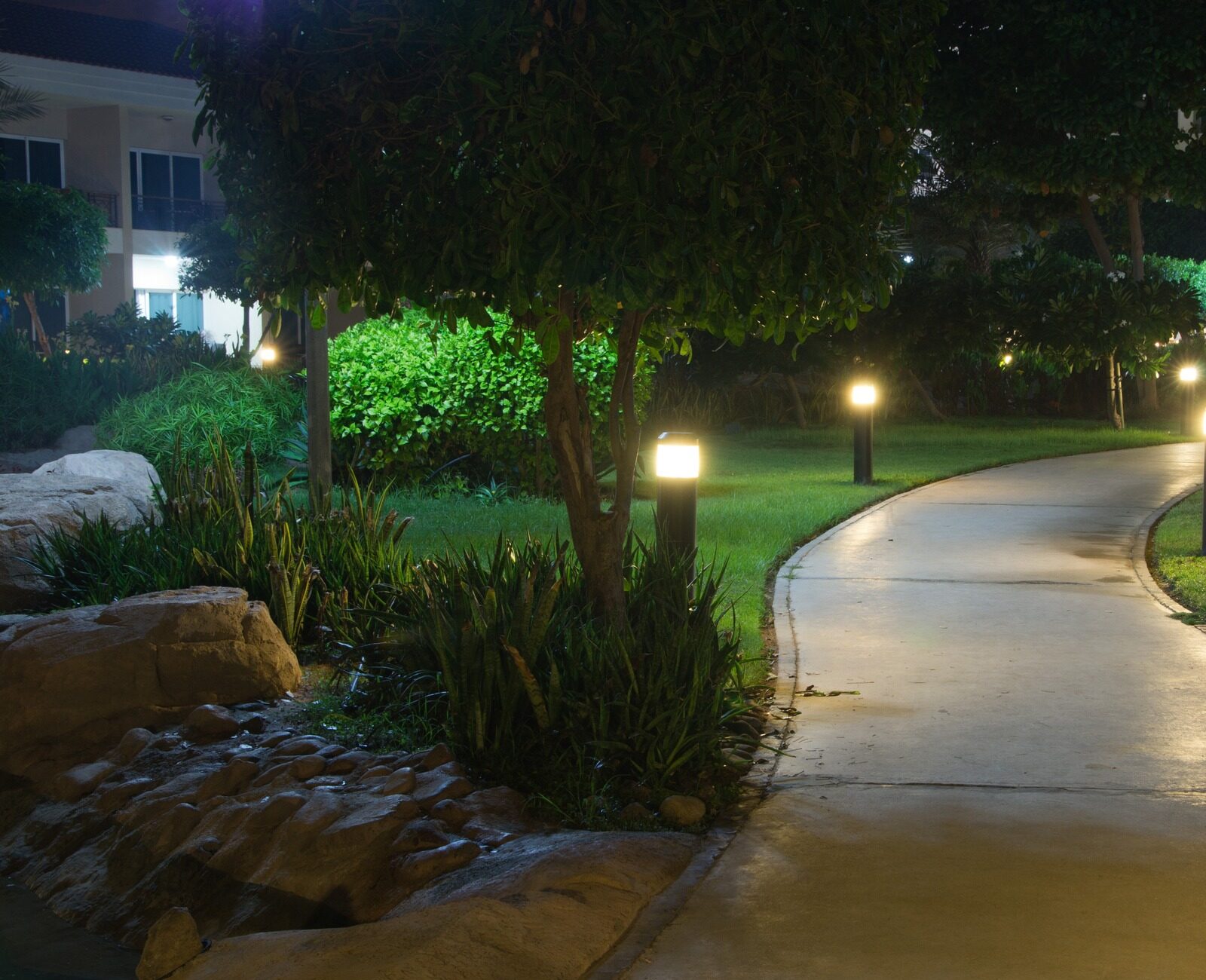 Illuminated garden path at night, surrounded by lush greenery and rocks, with buildings visible in the background, creating a serene atmosphere.