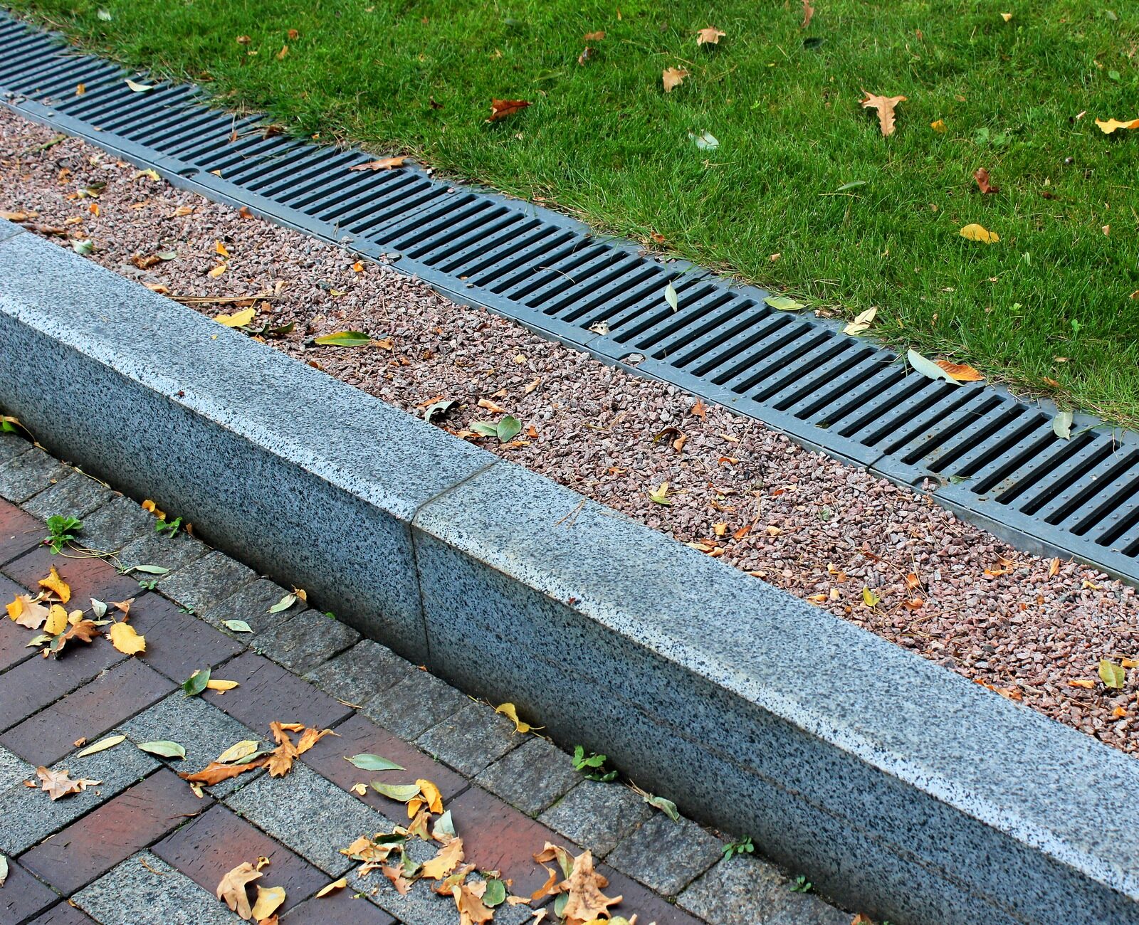 Grass verge with metal drainage grate, adjacent to a stone curb-lined path scattered with autumn leaves on a brick pavement.