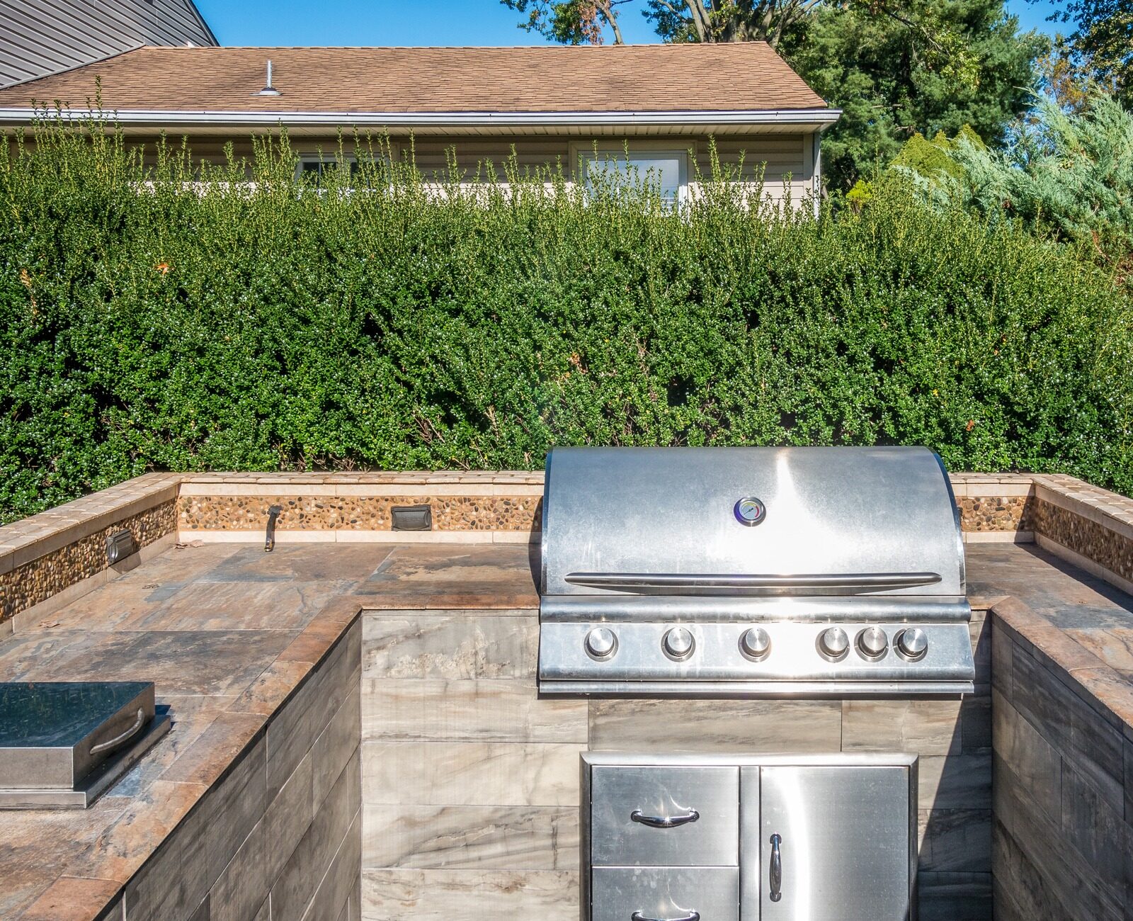 An outdoor kitchen features a sleek stainless steel grill set in a stone countertop, surrounded by lush green foliage and privacy fencing.