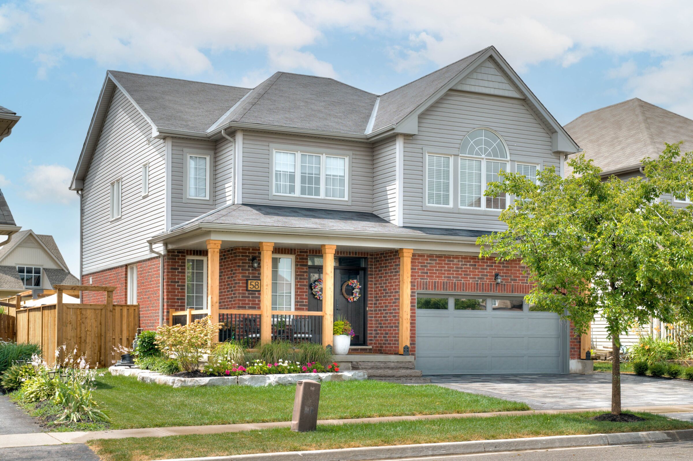 A two-story suburban house with brick and siding exterior, a front porch, a garage, and a well-maintained lawn with flowers and trees.