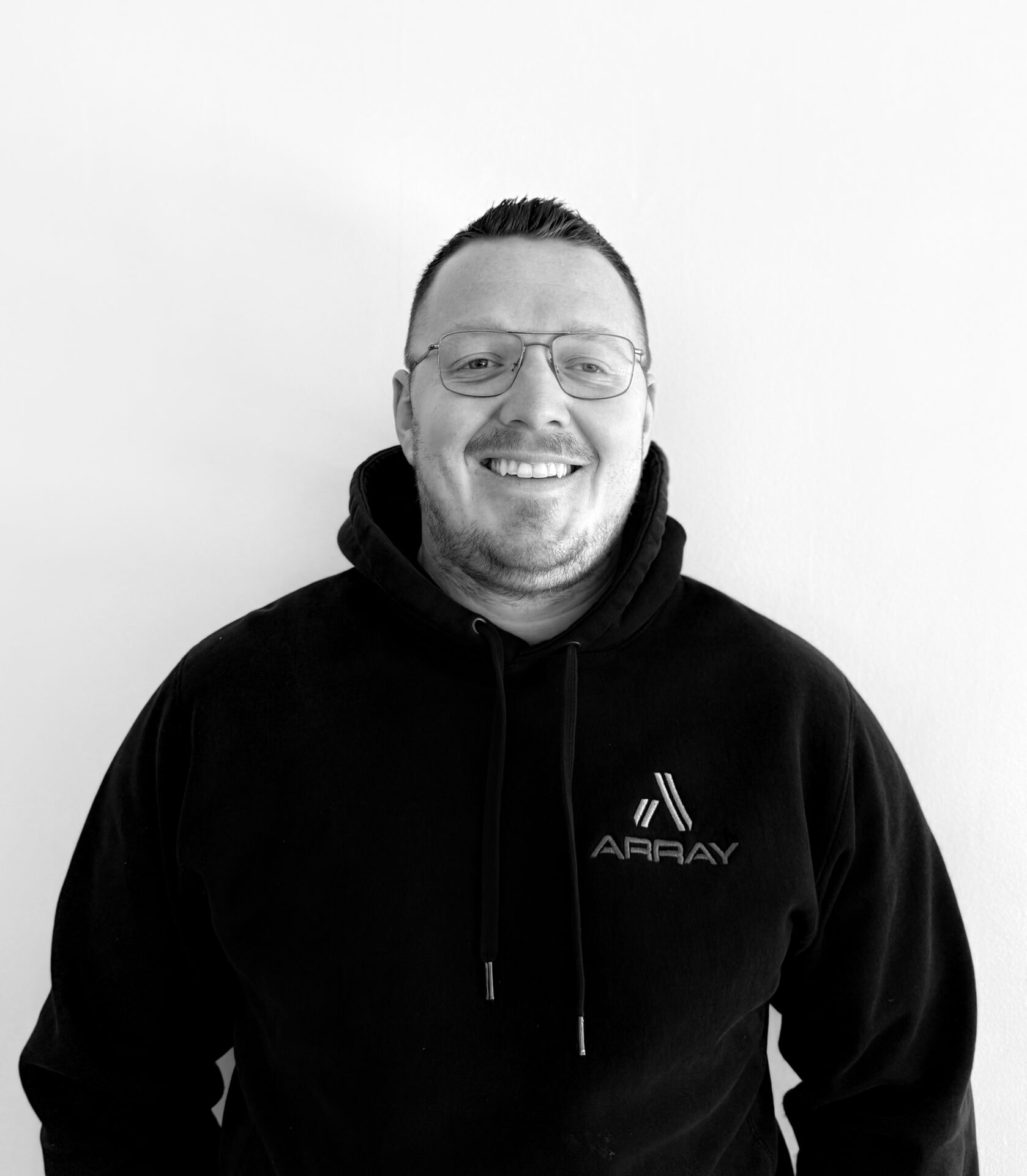A smiling person wearing glasses and a black "ARRAY" hoodie stands against a plain white background. No landmarks or historical buildings visible.